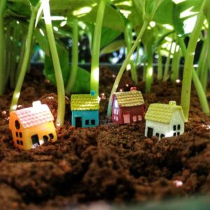 This is an image of 4 Miniature Resin House of different colors kept on soil with small plants on background.